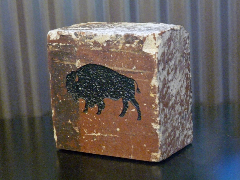 May include: A weathered, rectangular block with a brown base and a black bison silhouette. The block has a distressed, aged appearance with white paint chipping off the edges, giving it a rustic aesthetic.
