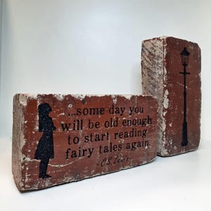 May include: Two weathered red bricks with black graphics and text. One brick features a silhouette of a girl and the quote "...some day you will be old enough to start reading fairy tales again." The other brick has a street lamp.