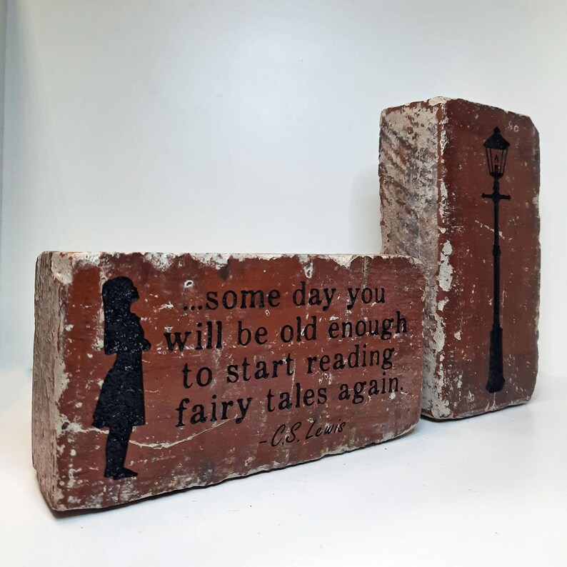 May include: Two weathered red bricks with black graphics and text. One brick features a silhouette of a girl and the quote "...some day you will be old enough to start reading fairy tales again." The other brick has a street lamp.