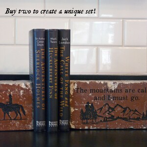K&ouml;nnte beinhalten: Ziegelstein-Buchst&uuml;tzen mit schwarzen Illustrationen und Text. Eine zeigt einen Reiter auf einem Pferd, die andere eine Berglandschaft mit dem Text "The mountains are calling and I must go." Drei klassische B&uuml;cher stehen dazwischen.