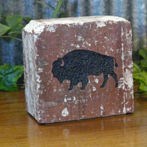 May include: A rustic, rectangular block with a distressed, reddish-brown surface and white paint accents. A black silhouette of a bison is featured on the front. The block is likely made of wood and has a weathered appearance.