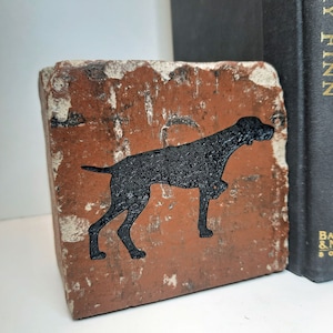 May include: A rustic, square brick bookend featuring a black silhouette of a dog. The brick has a weathered, reddish-brown appearance with white accents. The bookend is positioned next to a stack of black books with gold lettering.