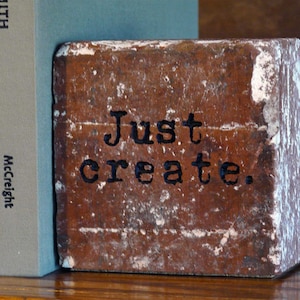 May include: A weathered, rectangular brick with the words "Just create." in black lettering. The brick is brown with white paint chipping off the edges. It is positioned between two books on a wooden surface.