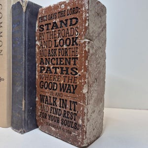 May include: A rectangular, weathered brick with the text "THUS SAYS THE LORD: STAND BY THE ROADS AND LOOK AND ASK FOR THE ANCIENT PATHS WHERE THE GOOD WAY IS; AND WALK IN IT AND FIND REST FOR YOUR SOULS." The brick is brown with black lettering.