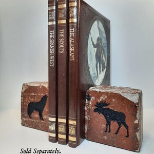 Op de afbeelding: Een verzameling vintage boeken getiteld "The Spanish West", "The Scouts" en "The Alaskans" met bruine covers. De boeken worden bij elkaar gehouden door twee bakstenen boekensteunen, &eacute;&eacute;n met een zwart wolfsilhouet en de andere met een zwart elandsilhouet.
