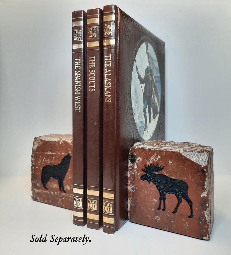 Op de afbeelding: Een verzameling vintage boeken getiteld "The Spanish West", "The Scouts" en "The Alaskans" met bruine covers. De boeken worden bij elkaar gehouden door twee bakstenen boekensteunen, &eacute;&eacute;n met een zwart wolfsilhouet en de andere met een zwart elandsilhouet.