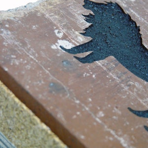 May include: A weathered, rectangular brick with a dark, silhouetted bird design. The bird is painted in black against the reddish-brown brick. The brick has a rough, textured surface with white speckles.