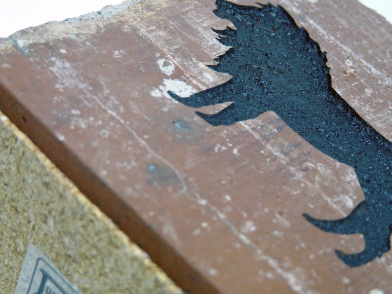 May include: A weathered, rectangular brick with a dark, silhouetted bird design. The bird is painted in black against the reddish-brown brick. The brick has a rough, textured surface with white speckles.