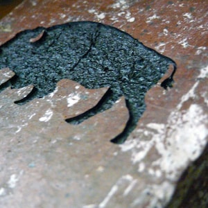 May include: A close-up of a rustic, rectangular stone with a carved silhouette of a bison. The bison is black and textured, contrasting with the reddish-brown and white speckled stone. The image is well-lit, highlighting the details of the carving.