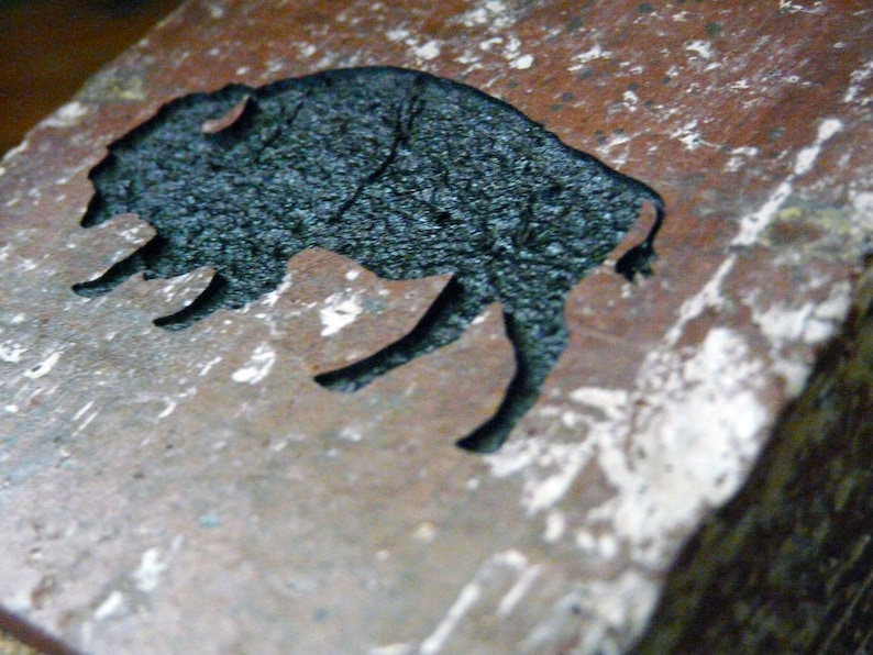May include: A close-up of a rustic, rectangular stone with a carved silhouette of a bison. The bison is black and textured, contrasting with the reddish-brown and white speckled stone. The image is well-lit, highlighting the details of the carving.