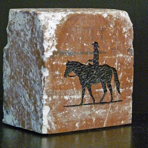 K&ouml;nnte beinhalten: Rechteckiger, rotbrauner Steinblock mit einer schwarzen Silhouette einer Person auf einem Pferd. Der Stein hat eine raue, strukturierte Oberfl&auml;che mit wei&szlig;en Flecken. Der Reiter tr&auml;gt einen Hut.