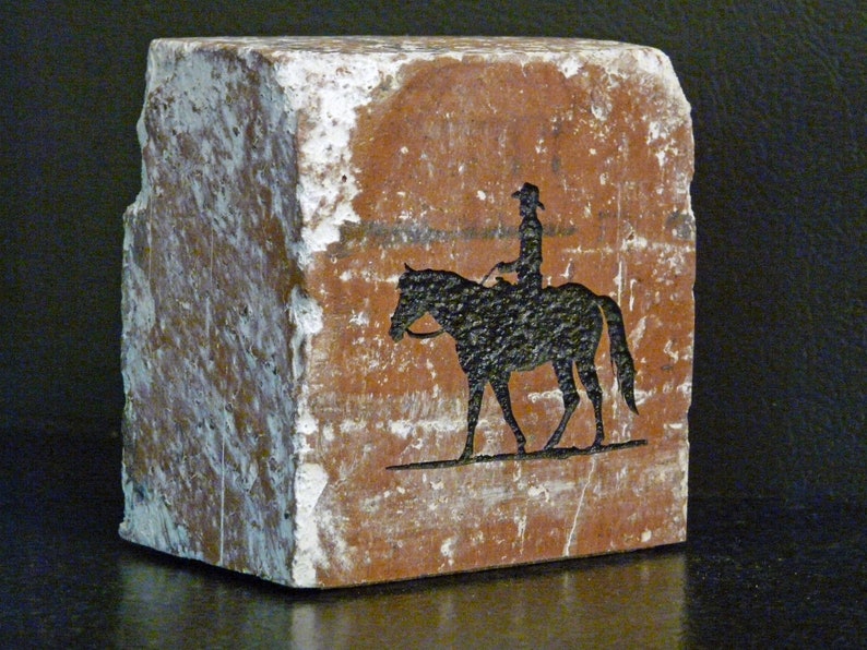 K&ouml;nnte beinhalten: Rechteckiger, rotbrauner Steinblock mit einer schwarzen Silhouette einer Person auf einem Pferd. Der Stein hat eine raue, strukturierte Oberfl&auml;che mit wei&szlig;en Flecken. Der Reiter tr&auml;gt einen Hut.