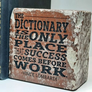 May include: A rectangular, brick-like decorative block with the quote "THE DICTIONARY IS THE ONLY PLACE THAT SUCCESS COMES BEFORE WORK" in black lettering. The block has a distressed, aged appearance with peeling white paint. Two dark blue books are visible in the background.