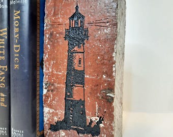 May include: A weathered, rectangular bookend with a black lighthouse design on a reddish-brown surface. The bookend is made of a textured material. Two dark blue books are visible behind the bookend, with the titles "Moby-Dick" and "White Fang and" visible.