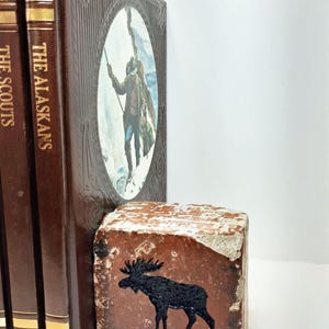 Op de afbeelding: Een verzameling vintage boeken met bruine covers en gouden letters, waaronder "The Alaskans" en "The Scouts". Een rustieke, rechthoekige bakstenen boekensteun met een zwart elandsilhouet staat voor de boeken. De boekensteun heeft een verweerde, verouderde uitstraling.