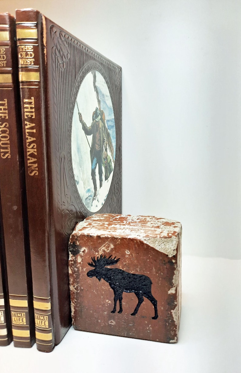 Op de afbeelding: Een verzameling vintage boeken met bruine covers en gouden letters, waaronder "The Alaskans" en "The Scouts". Een rustieke, rechthoekige bakstenen boekensteun met een zwart elandsilhouet staat voor de boeken. De boekensteun heeft een verweerde, verouderde uitstraling.