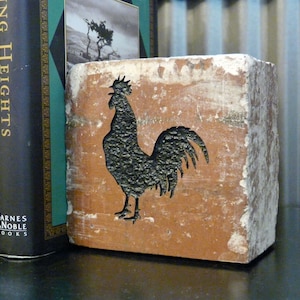 May include: A rustic, rectangular bookend with a rooster silhouette cut-out. The bookend is made of a weathered, reddish-brown material with a textured surface. The rooster is black. Two books are visible.