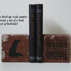 Puede incluir: Dos sujetalibros de ladrillo con dise&ntilde;os y texto pintados en negro. Un ladrillo presenta una silueta de lobo, el otro dice "IN THE WOODS IS PERPETUAL YOUTH". Dos libros negros se interponen entre ellos.