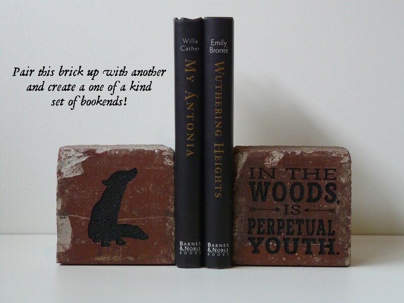 Puede incluir: Dos sujetalibros de ladrillo con dise&ntilde;os y texto pintados en negro. Un ladrillo presenta una silueta de lobo, el otro dice "IN THE WOODS IS PERPETUAL YOUTH". Dos libros negros se interponen entre ellos.