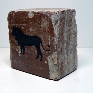 May include: A weathered, rectangular block with a rough, textured surface. The block is primarily brown with areas of lighter color. A black silhouette of a lion is painted on one side, adding a decorative element.