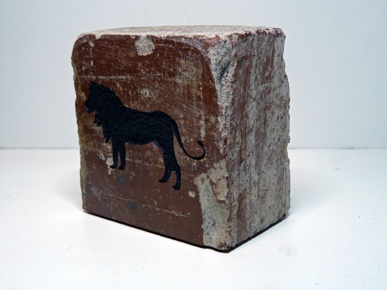May include: A weathered, rectangular block with a rough, textured surface. The block is primarily brown with areas of lighter color. A black silhouette of a lion is painted on one side, adding a decorative element.