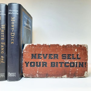 May include: A weathered, rectangular brick with the text "NEVER SELL YOUR BITCOIN!" in black lettering. The brick is reddish-brown and sits next to two dark blue hardcover books.