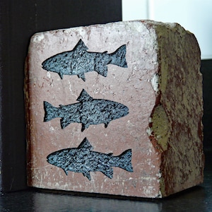 May include: A rustic, rectangular bookend made of brick, featuring three stylized black fish cutouts. The brick has a weathered, reddish-brown appearance with white and yellow paint accents, adding to its vintage charm. The bookend is positioned next to a stack of dark-colored books.