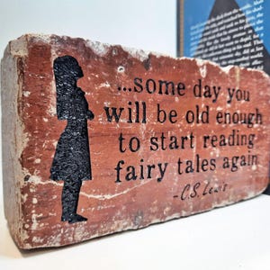 May include: A brick with a quote from C.S. Lewis, "...some day you will be old enough to start reading fairy tales again", alongside a black silhouette. The brick is reddish-brown, with a book in the background.