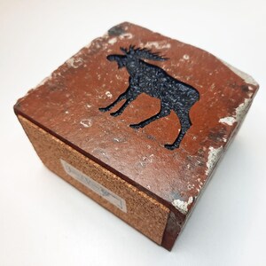 Op de afbeelding: Vierkante houten doos met een uitgesneden silhouet van een eland. De eland is zwart op een roodbruine achtergrond. De doos heeft een kurken basis en verweerde randen. Afmetingen: ongeveer 10 cm x 10 cm.