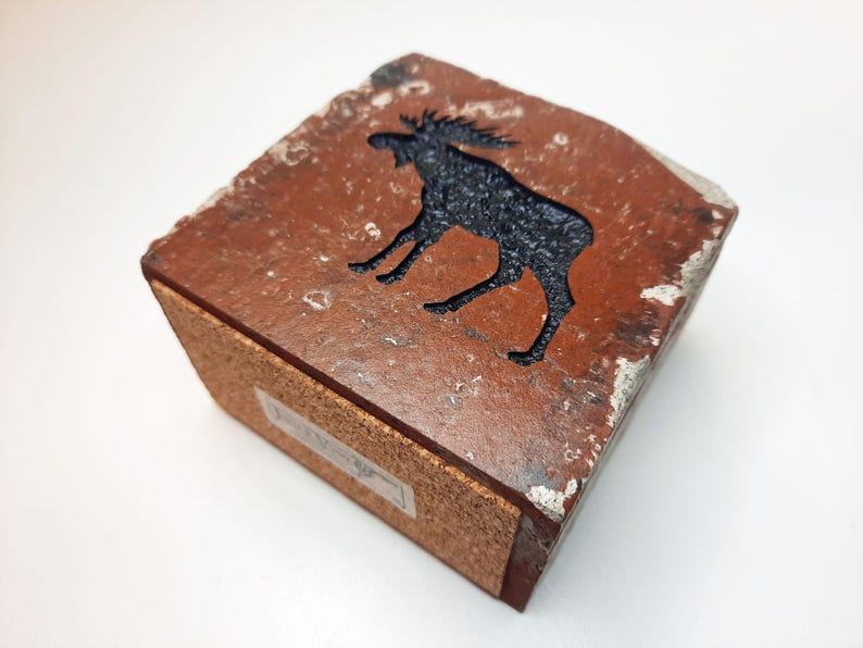 Op de afbeelding: Vierkante houten doos met een uitgesneden silhouet van een eland. De eland is zwart op een roodbruine achtergrond. De doos heeft een kurken basis en verweerde randen. Afmetingen: ongeveer 10 cm x 10 cm.