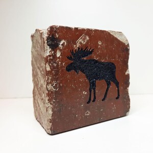 Op de afbeelding: Een rechthoekig blok van roodbruine steen met een zwart silhouet van een eland. De eland wordt in profiel weergegeven. De steen heeft een ruw, getextureerd oppervlak. De achtergrond is wit.