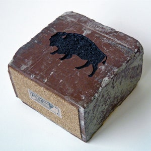 May include: A rectangular, brown block with a black bison silhouette on top. The bottom of the block is lined with cork. A small label is affixed to the cork. The block has a rough, textured appearance.