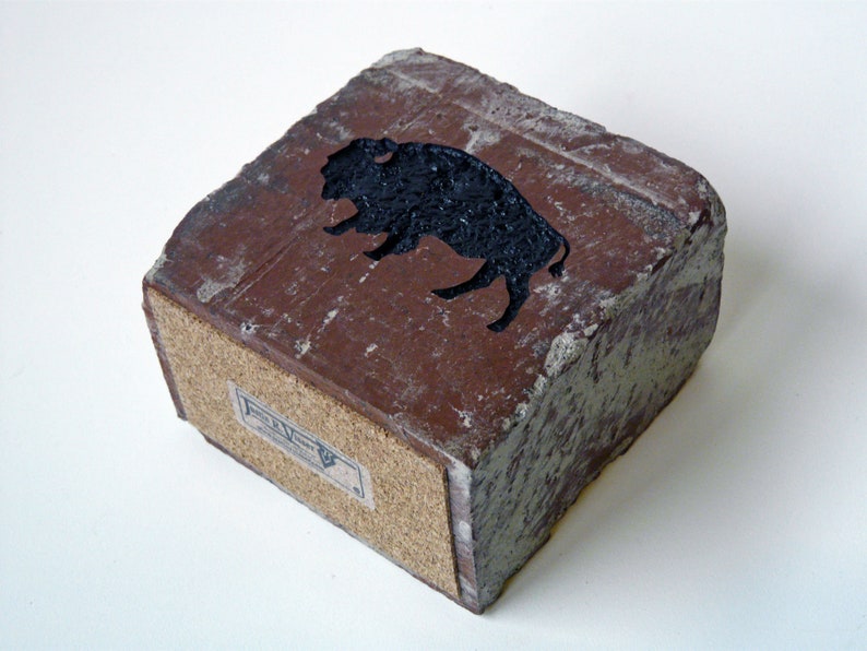 May include: A rectangular, brown block with a black bison silhouette on top. The bottom of the block is lined with cork. A small label is affixed to the cork. The block has a rough, textured appearance.