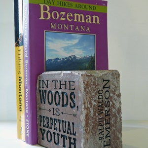 Puede incluir: Libros, incluyendo "Day Hikes Around Bozeman, Montana", sostenidos por un sujetalibros rectangular de piedra. El sujetalibros tiene la cita "In the woods is perpetual youth" de Ralph Waldo Emerson. Fondo blanco.