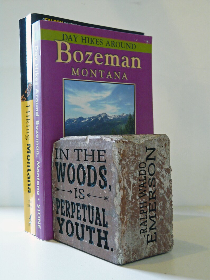 Puede incluir: Libros, incluyendo "Day Hikes Around Bozeman, Montana", sostenidos por un sujetalibros rectangular de piedra. El sujetalibros tiene la cita "In the woods is perpetual youth" de Ralph Waldo Emerson. Fondo blanco.