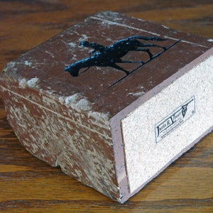 K&ouml;nnte beinhalten: Rechteckige, braune Steinbox mit einer schwarzen Silhouette eines Jockeys auf einem Pferd. Die Box hat einen Korkboden und ein abgenutztes, strukturiertes Aussehen. Die Box hat den Text "Jasmin R. Visser" auf der Unterseite.