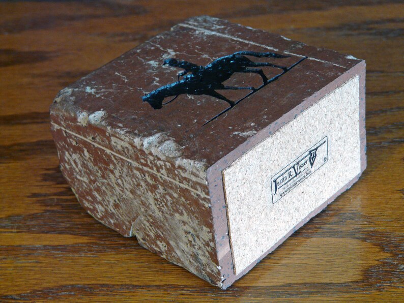 K&ouml;nnte beinhalten: Rechteckige, braune Steinbox mit einer schwarzen Silhouette eines Jockeys auf einem Pferd. Die Box hat einen Korkboden und ein abgenutztes, strukturiertes Aussehen. Die Box hat den Text "Jasmin R. Visser" auf der Unterseite.