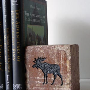 Op de afbeelding: Een rustieke bakstenen boekensteun met een zwart elandsilhouet staat naast drie hardcover boeken. De boeken heten "The Adventures of Huckleberry Finn" en "The Strange Case of Dr. Jekyll and Mr. Hyde".