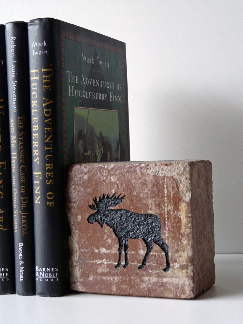 Op de afbeelding: Een rustieke bakstenen boekensteun met een zwart elandsilhouet staat naast drie hardcover boeken. De boeken heten "The Adventures of Huckleberry Finn" en "The Strange Case of Dr. Jekyll and Mr. Hyde".