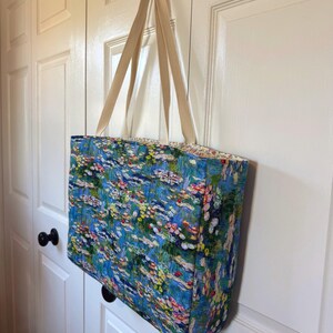 May include: A rectangular tote bag featuring a vibrant, multi-colored impressionist-style floral print with shades of blue, green, pink, and yellow. The bag has two long, cream-colored fabric straps and a quilted texture throughout the exterior.