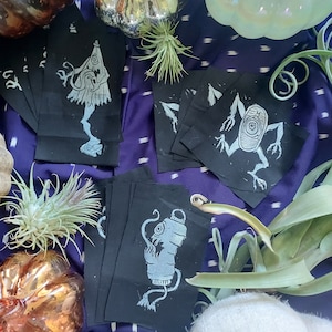 May include: A set of five black fabric patches with white ink illustrations of various spooky creatures. The patches feature a creature with a lantern, a creature with a cone head, and a creature with a large eye. The patches are surrounded by decorative pumpkins and plants.