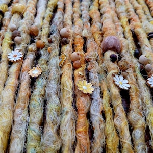 May include: Close-up of handmade dreadlocks in shades of yellow, orange, and brown, adorned with wooden beads and daisy-shaped charms. The dreadlocks are arranged vertically, creating a textured and colorful display.