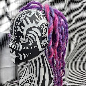 May include: A black and white painted mannequin head with a purple and pink braided hair extension. The mannequin is painted with abstract patterns and designs.