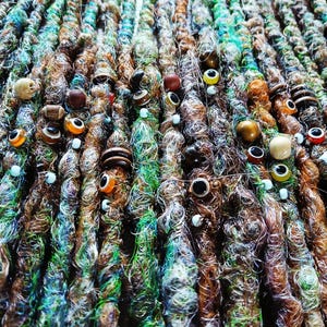 May include: Close-up of handmade dreadlocks in various shades of brown, green, and blue. The dreads are adorned with beads, wooden accents, and googly eyes. The texture is intricate and detailed, showcasing a unique, artistic style.