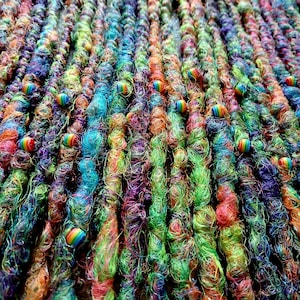 Puede incluir: Primer plano de rastas coloridas en tonos de azul, verde, morado y naranja. Cada rasta está adornada con una pequeña cuenta de arcoíris. La textura del cabello es visible, con mechones de varios colores entrelazados.