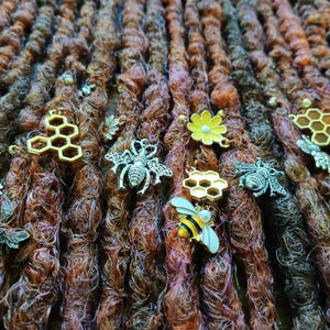 Könnte beinhalten: Nahaufnahme von braunen und kastanienbraunen Dreadlocks, die mit verschiedenen Charms zum Thema Bienen verziert sind. Zu den Charms gehören goldene Wabenformen, silberne Bienen und gelbe und weiße Blumenakzente.
