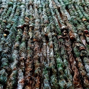 May include: Close-up of handmade dreadlocks in shades of green, brown, and gray. The dreads are adorned with small wooden beads and metal leaf charms, creating a natural, earthy aesthetic. The texture is rough and detailed.
