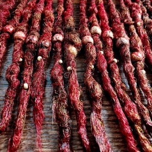 May include: Close-up of several red and brown dreadlocks, adorned with small, round, off-white beads. The dreadlocks are arranged closely together, creating a textured, organic pattern. The background is a dark, wooden surface.