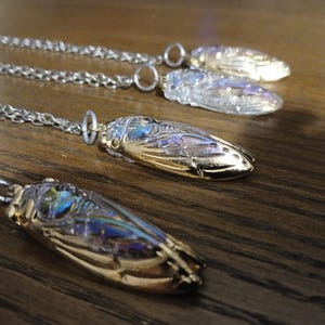 Può includere: Quattro ciondoli a forma di insetto su catene d'argento. I ciondoli presentano dettagli iridescenti, trasparenti e dorati. I gioielli sono esposti su una superficie di legno scuro. I ciondoli sono probabilmente collane.