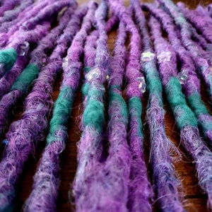 Pode incluir: Close-up de um conjunto de dreadlocks sintéticas roxas e verde-azuladas com contas transparentes. As dreadlocks estão dispostas em filas e estão prontas para serem instaladas.